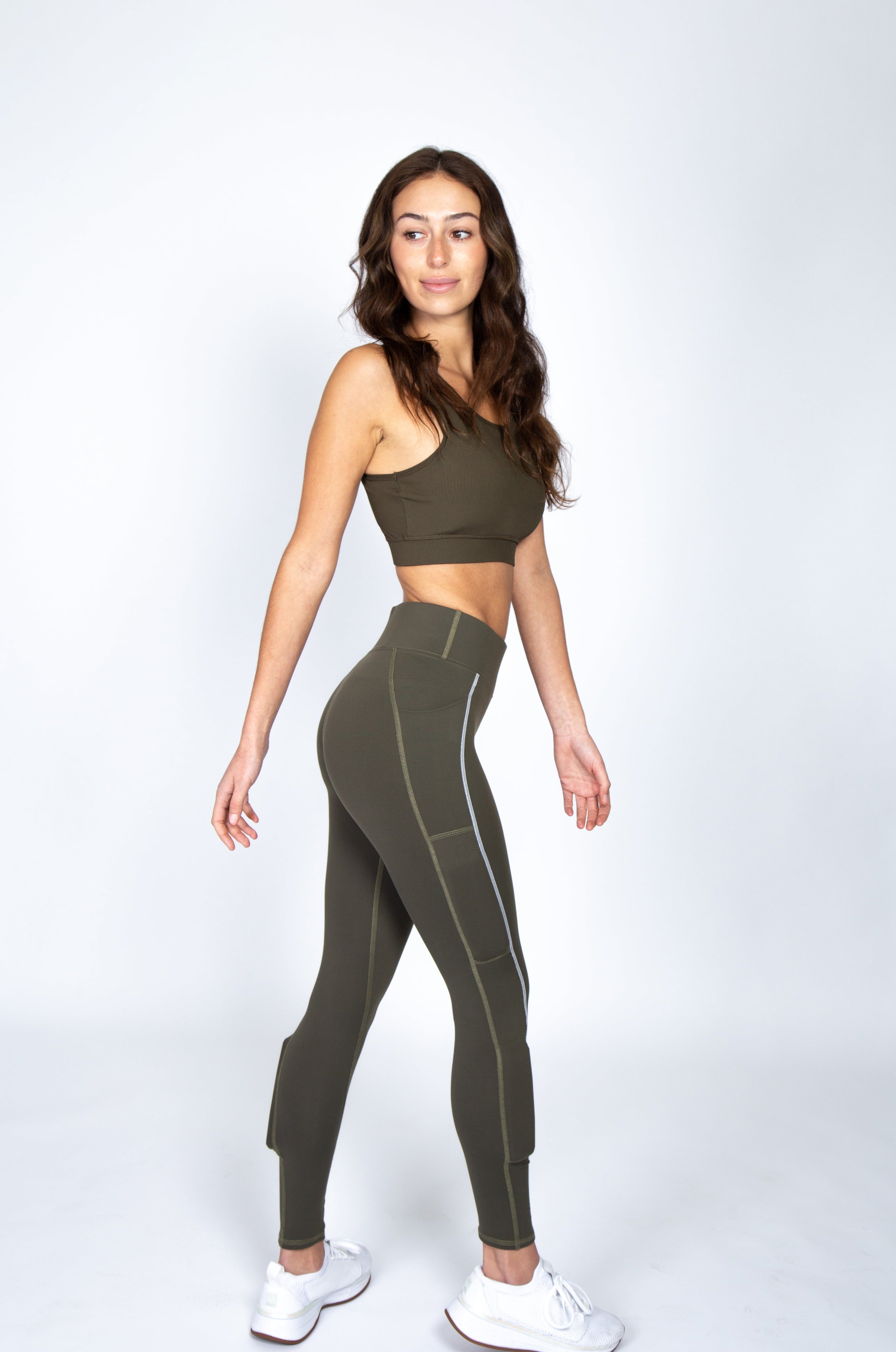 Woman wearing army green bra, turning to the side in the the army green legging called juniper lift with white stitch that runs down the leg,  weight in the weight pocket above the knee and weight in the below the knee on the side calf.