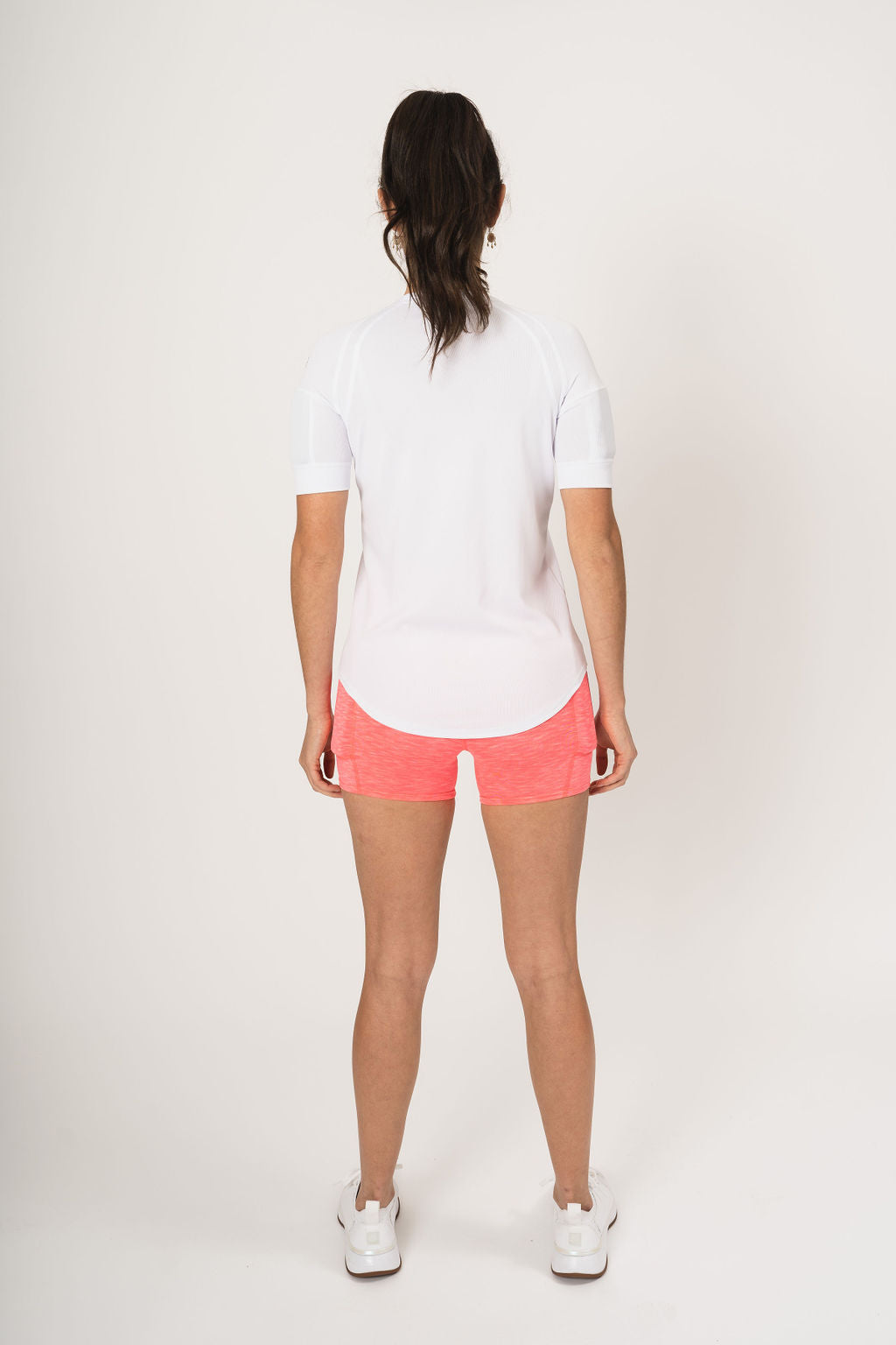 rear view showing woman wearing the weighted white short sleeve with weights on the biceps and the heather coral shorts with weights in the side weight pockets, found on the quads.
