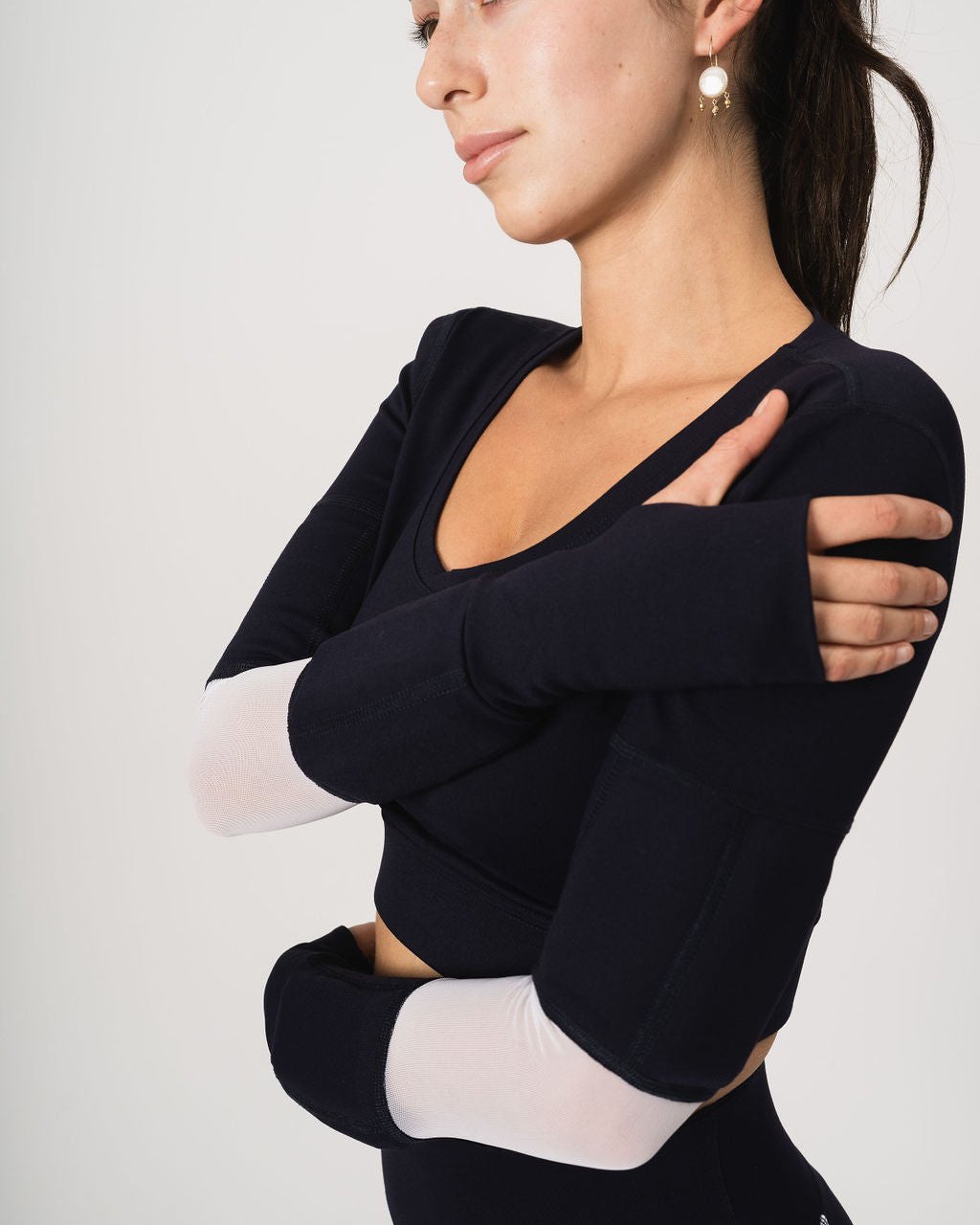 Woman wearing scoop neck long sleeve crop top in navy with white mesh at the elbow. She is wearing two weights in the bicep weight pockets and two weights above the wrist on the forearms. She has her arms folded across her body.