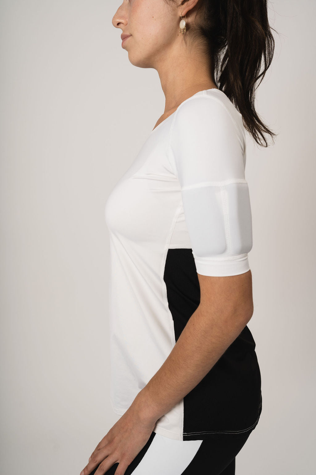 Close up Side view woman wearing the relaxed fit weighted short sleeve. Top is all white in the front with a scoop neck. The back has white at the top and black mesh halfway down the back. Weights are in the bicep arm pockets.