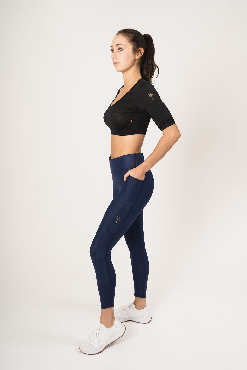 Woman wearing the weighted black crop tops with the weights on the arms and wearing the navy shiny leggings with the weights down the legs, with one weight above the knee and one weight below the knee and hands in the cell phone pocket