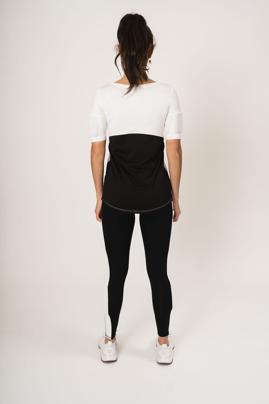 Rear view woman wearing the relaxed fit weighted short sleeve. The back has white at the top and black mesh halfway down the back. Weights are in the bicep arm pockets.