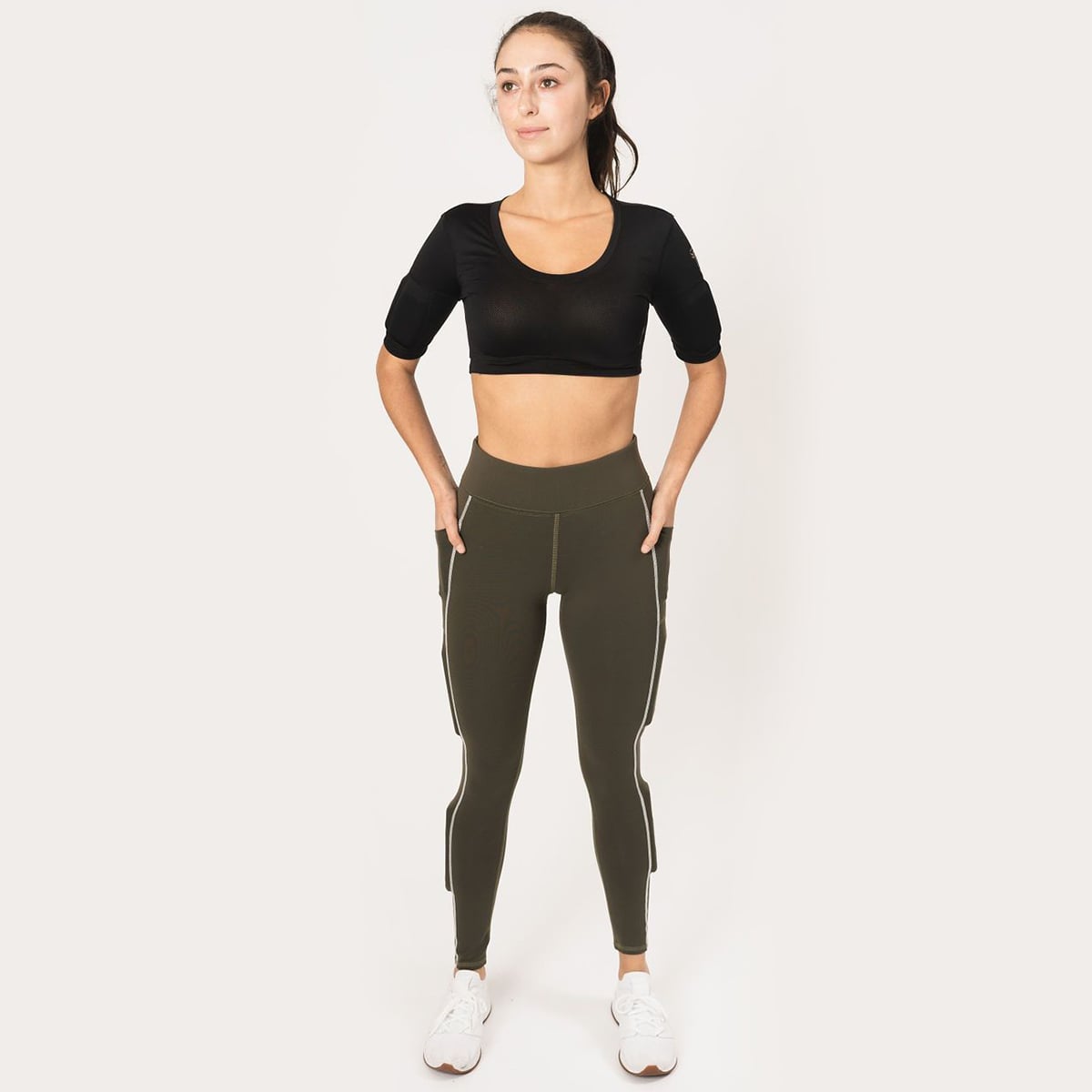 Woman wearing black weighted crop top, front view, in the the army green legging called juniper lift, hands in side cell phone pockets, with white stitch that runs down the leg, weight in the weight pocket above the knee and weight in the below the knee on the side calf.